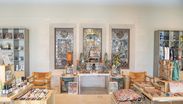 Photograph of museum shop at The Burrell Collection. The room has ambient orange tones as it is well lit from the sun. There are many items on display in front of stained glass windows which are not lit from outside light. You can see books, vases clothing and other items.