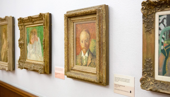 angled view of oil paintings in golden frames on a white wall, the focal image is a Van Gogh painting of a man with a ginger beard, face on.
