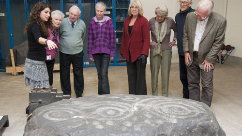 A group of visitors take part in a tour of a museum store. They are looking at an ancient stone carved with decorative shapes.