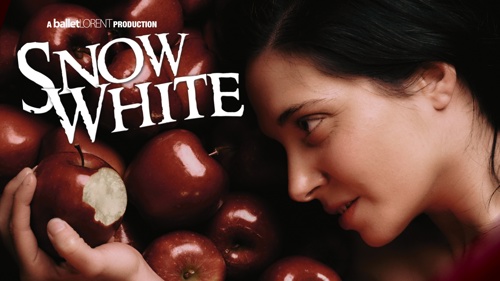 Close up of person with long dark hair, lying on a pile of red shiny apples. They are holding one of the apples in front of them. It has a large bite taken out of it