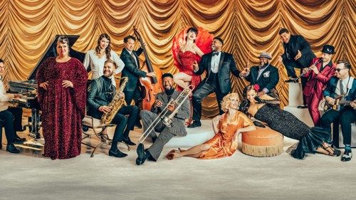 A large group of performers and musicians wearing glamorous 1920s clothing are gathered on a stage with a gold backdrop