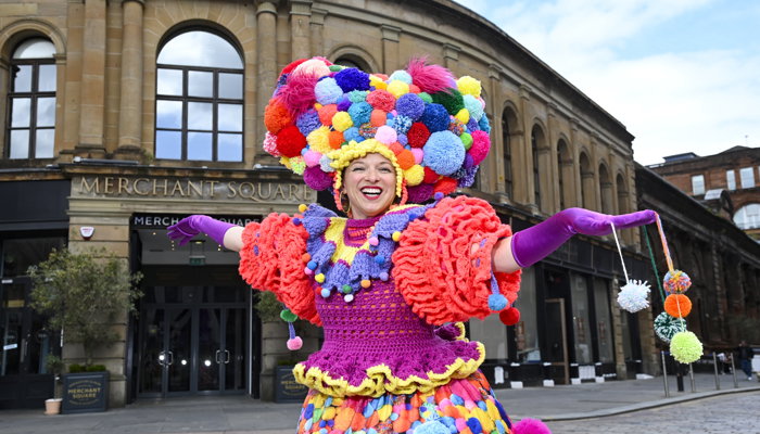 A performer in a brightly coloured outfit made of pom-poms stands in front of a 1700s building. A sign for Merchant Square can be seen behind the performer.