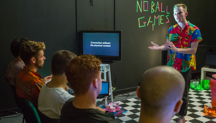 A person, is performing a play they are wearing a tropical shirt and pointing to a TV. In the background is neon tape that reads "No Ball Games".