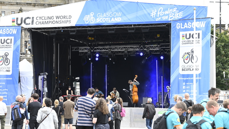 Musicians performing on the main stage in the George Square fan zone during the 2023 UCI Cycling World Championships. Two are sitting playing guitar while one is standing playing double bass and another the keyboard.