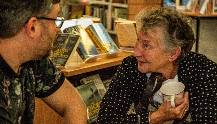 A adult and an elderly person sit in a  public library chatting. 