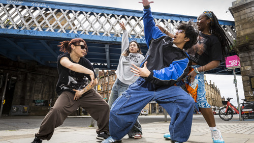 A group of four hip hop dancers are mid-move dancing outside in the city centre near a blue railway bridge and sandstone tenements 