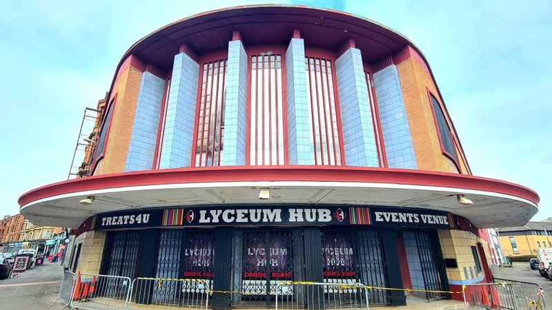 A curved, art deco-style building with tall vertical windows and a red-trimmed roof stands on a street corner. The front signage reads “LYCEUM HUB”. Metal barriers are placed along the pavement in front of the closed entrance, and surrounding buildings and a cloudy blue sky are visible in the background.