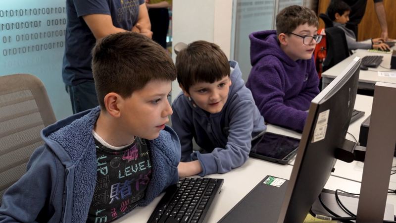 Three young people use desktop computers during a coding workshop in a library.