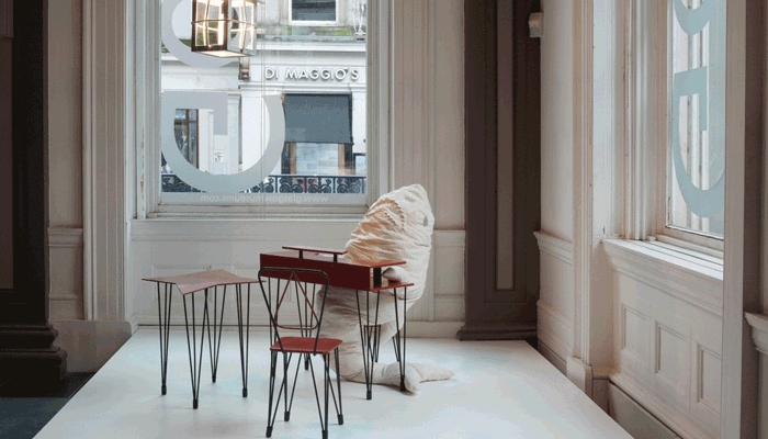 A staged gallery corner with small red metal tables and chairs by a tall window, where a soft, white sculpture of shark is seated on one of the chairs at red metal desk.