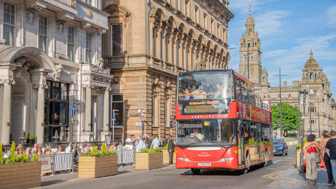  A red double decker bus is driving on a road, people are sitting at tables enjoying food and drink, there is sandstone buildings and blue skies behind the bus