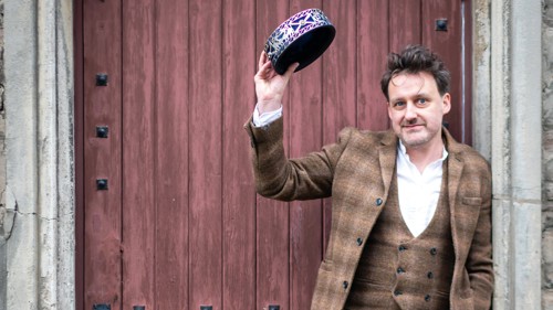 A person wearing a Tweed jacket and waistcoat, is standing in a stone doorway in front of an old wooden door. 