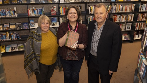 Three people gathered for a group photo in a library. The person in the middle is holding up a book