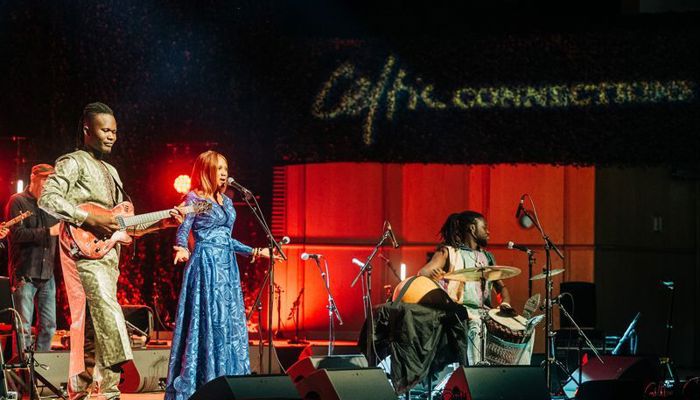 A musical group performs on a stage lit with red and gold lights under a banner for "Celtic Connections." A woman in a vibrant blue patterned dress sings into a microphone at center stage. To her left, a man in a shimmering gold outfit plays an electric guitar. To her right, a percussionist plays a djembe and other drums. The atmosphere is energetic, with stage monitors and microphones visible in the foreground.