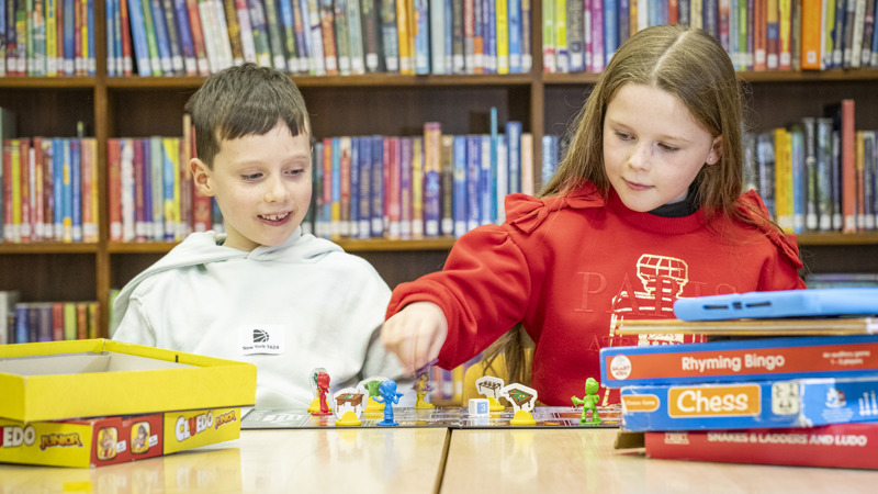 Two children playing board games on a wooden table in front of shelves of books in a library