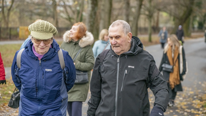 Two older people walking the park during a health walk, both are wearing jackets and one has a khaki green hat on.