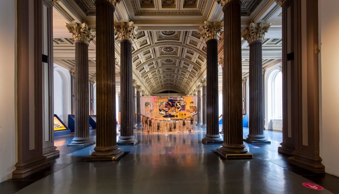 A grand, ornate gallery hall with tall columns and a vaulted ceiling, featuring a colourful large tapestry suspended between the main aisle.