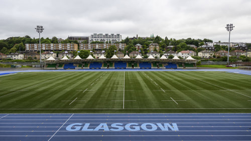 A blue and white running track surrounds a rugby pitch and a stand and housing are visible in the background