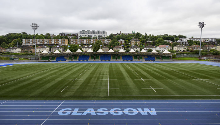 A blue and white running track surrounds a rugby pitch and a stand and housing are visible in the background