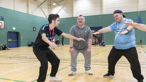  Three men smiling and participating in an activity in a brightly lit indoor sports hall.