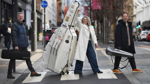 Three people walk across a street carrying musical instruments