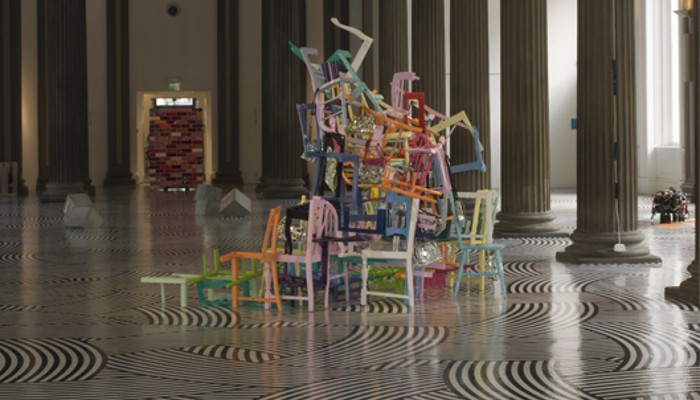 A tall, ornate gallery hall with decorative columns and a patterned floor, featuring a central sculpture made of stacked, colourful chairs and suspended abstract forms hanging from the ceiling above.