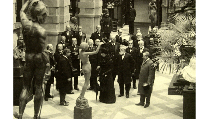 black and white image of men in black suits in a gallery setting looking at a marble statue of a woman 