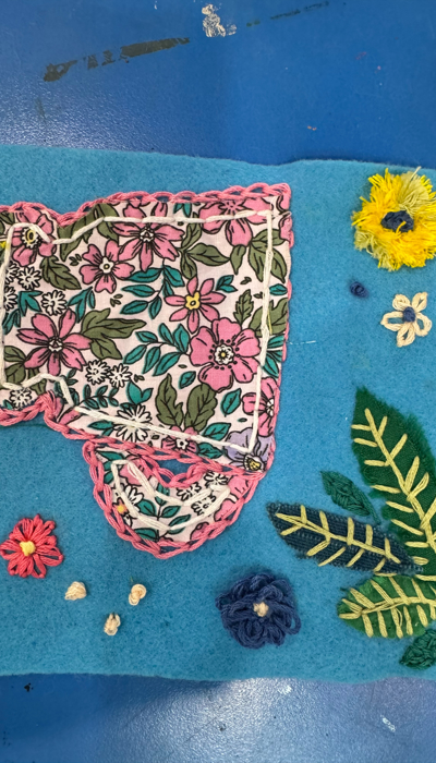 A piece of blue felt decorated with appliqué and embroidery. At the centre is a floral-patterned fabric cut into an irregular outline and bordered with pink stitching. Around it are embroidered flowers in yellow, red, white, and dark blue, along with green felt leaves detailed with stitched veins. The background shows a blue work surface with paint marks and a strip of dark red felt along the right edge.