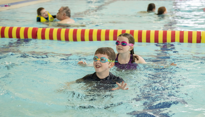Two children wearing swimming goggles play in an indoor pool, partially submerged in clear blue water. A red-and-yellow lane divider floats behind them, and other swimmers are visible in the background.