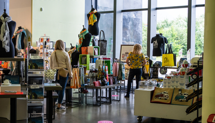 A store interior with various items on display, including bags, scarves, books, and other merchandise. Two people are browsing the items. The store has large windows allowing natural light to enter.