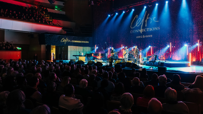 Audience seated in a concert hall watching a live band perform on a brightly lit stage under blue and red spotlights.