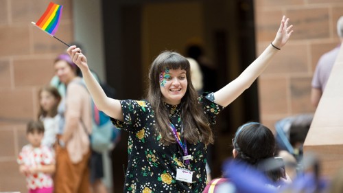 A person with arms outstretched is waving a small rainbow flag while leading a group activity for young people in a large sandstone museum hall