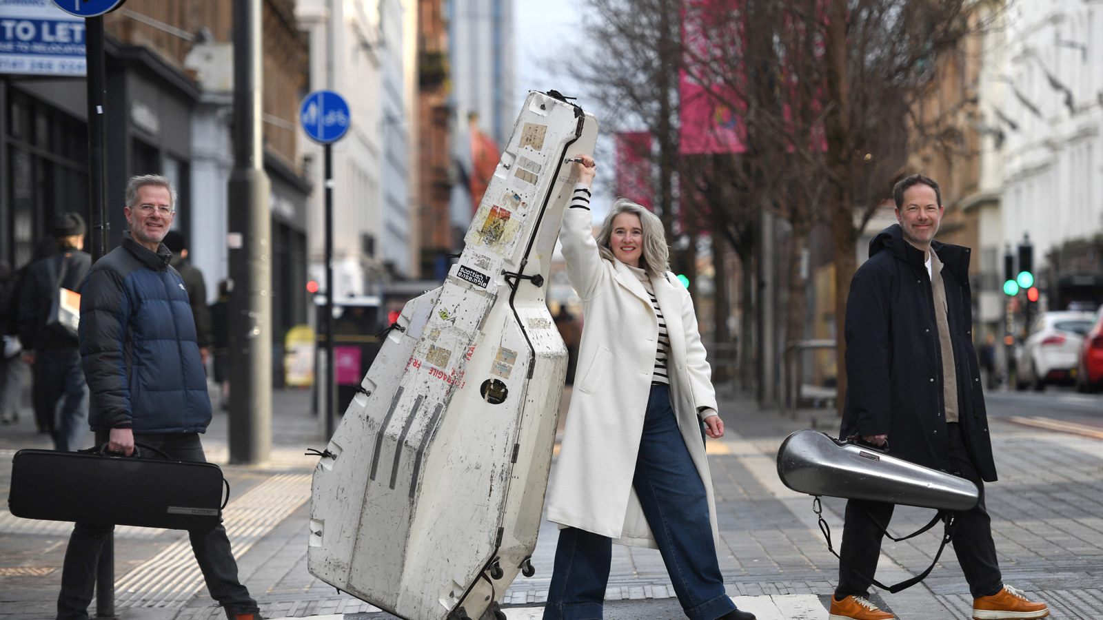 A group of three people carrying large musical instrument cases walking across a city centre pedestrian crossing, with tall buildings, street signs, and traffic in the background.