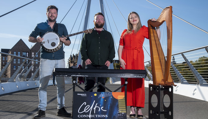 Three young musicians standing on the Govan-Partick bridge, one is holding a banjo, another is in front of a piano and the third is next to a harp.
