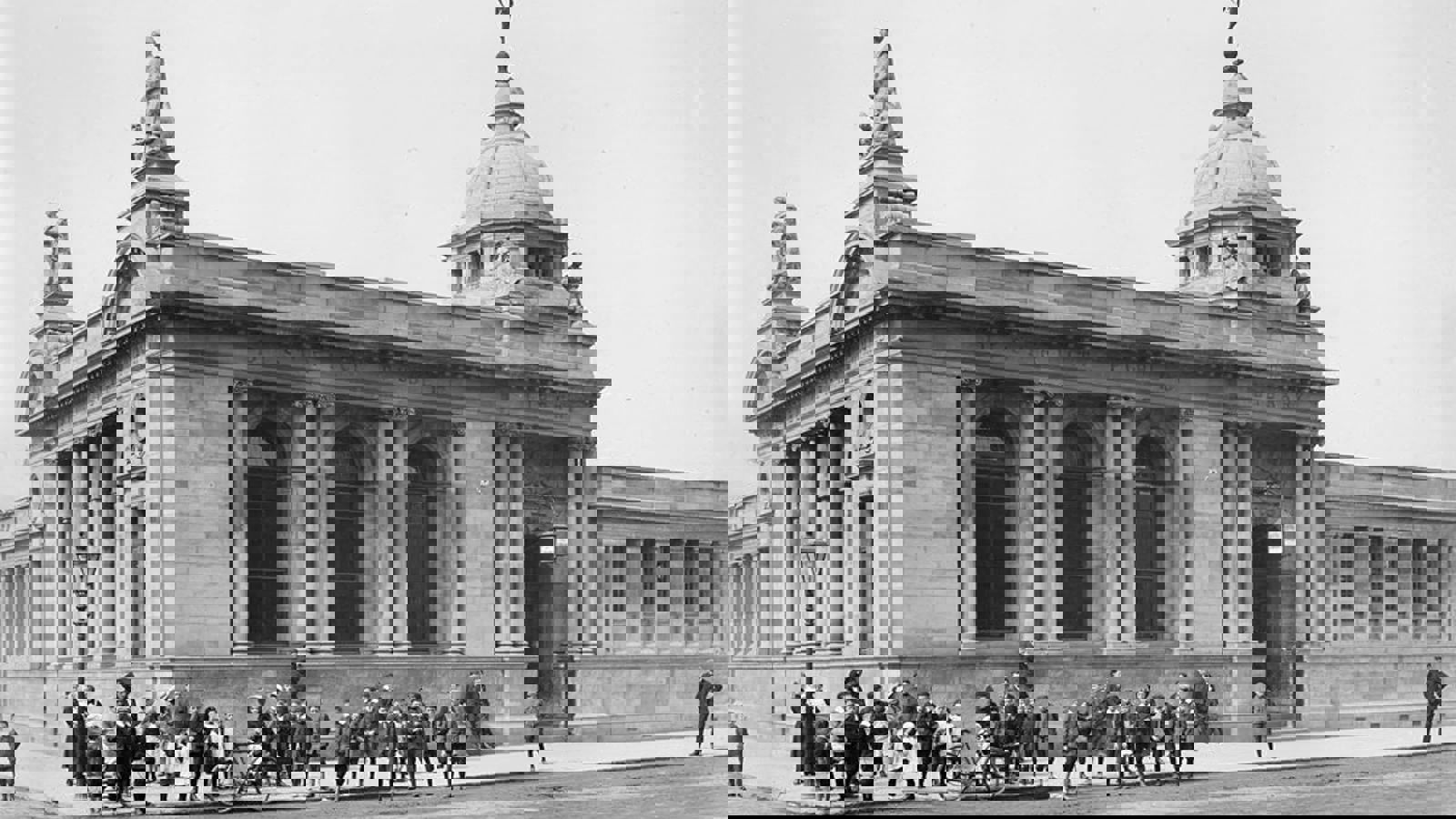 A black and white photograph of Govanhill Library which architecture includes pillars, arches and statues. Many people are on the street outside including adults, children and a bike.