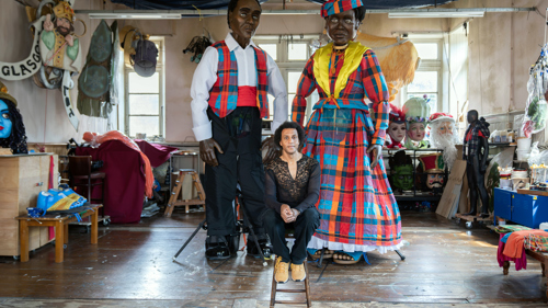 A person sits on a stool inside an artist's workshop between two towering five-metre puppets dressed in brightly coloured Madras-patterned costumes inspired by the Caribbean Wob Dwiyet tradition. The large-scale figures, one wearing a tartan-style waistcoat and the other a flowing checked dress with elaborate headwear, stand amid carnival masks, costume pieces and puppetry materials in the Glasgow studio where they were created for Merchant City Festival.