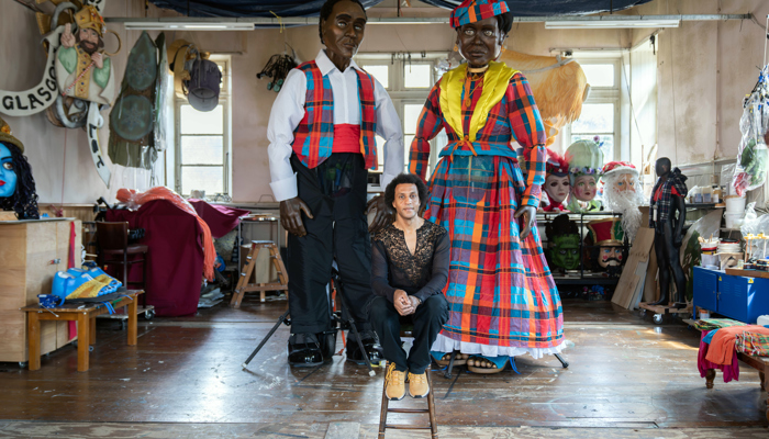 A person sits on a stool inside an artist's workshop between two towering five-metre puppets dressed in brightly coloured Madras-patterned costumes inspired by the Caribbean Wob Dwiyet tradition. The large-scale figures, one wearing a tartan-style waistcoat and the other a flowing checked dress with elaborate headwear, stand amid carnival masks, costume pieces and puppetry materials in the Glasgow studio where they were created for Merchant City Festival.