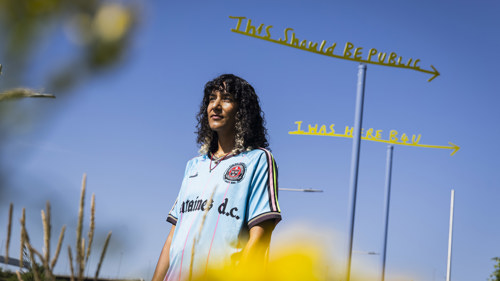 A woman wearing a blue top stands in front of a yellow sign set against a blue sky