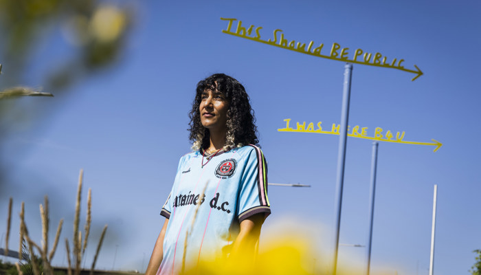 A woman wearing a blue top stands in front of a yellow sign set against a blue sky