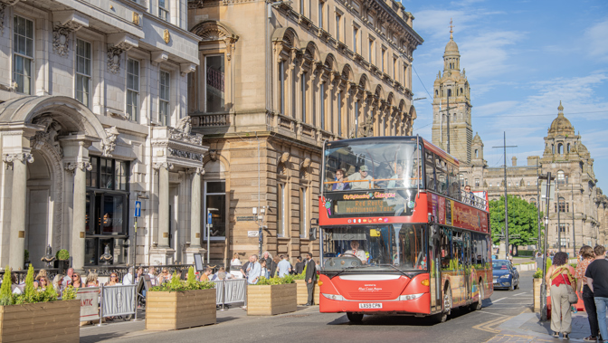 A red sightseeing bus is being driven along a road. There are old buildings in the background with a blue sky overhead
