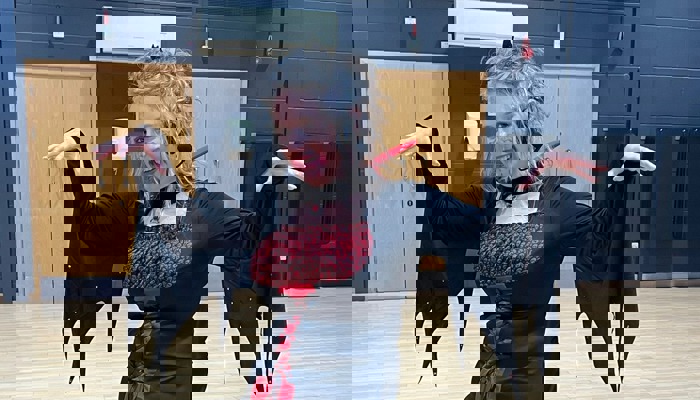 A person dressed in a red and black witches costume is standing with their arms outstretched in a sports hall