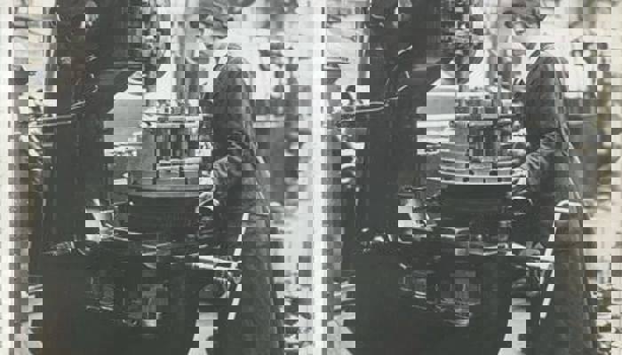 A black and white photo of an employee operating on a large slotting machine, wearing a long dark uniform with white collar and hat.