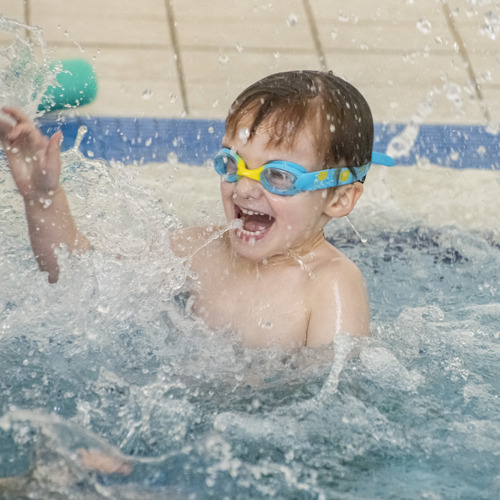 A young child splashing in a swimming pool they are smiling and wearing bright blue and yellow goggles