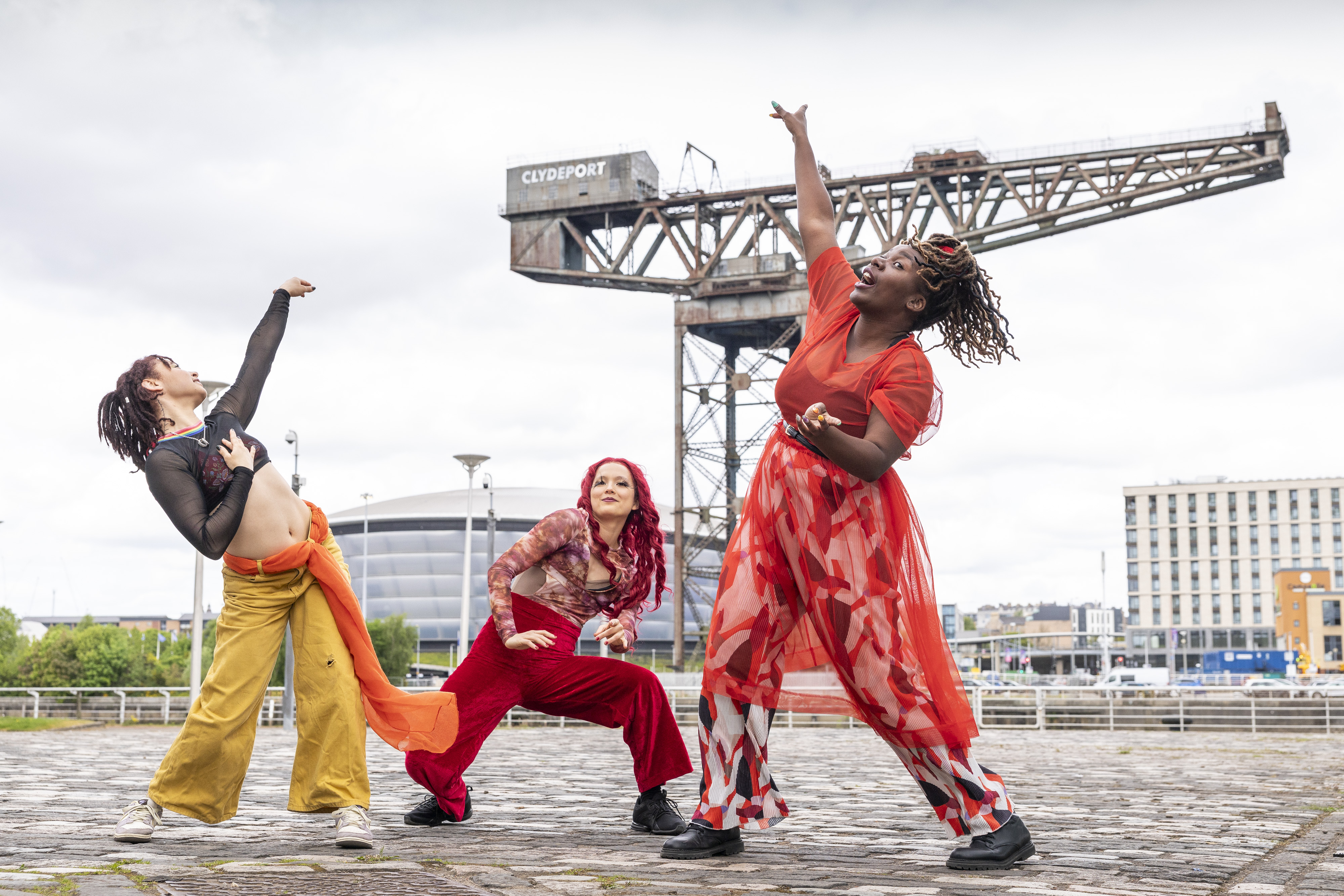 Three dancers from Whacking Scotland strike dynamic poses on a cobblestone plaza in Glasgow, with the Finnieston Crane and SEC Armadillo behind them. On the left, Nicolette McCormack leans back with an arm raised, wearing yellow trousers and a black mesh top. In the center, Leah James strikes a low stance in red velvet trousers, her long red hair down. On the right, Dorine Mwesiga reaches upward mid-motion, wearing a flowing, semi-transparent red dress over boldly patterned wide-leg pants.