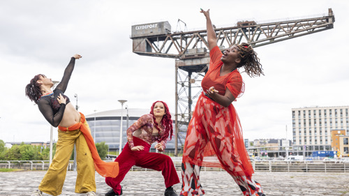 Three dancers from Whacking Scotland strike dynamic poses on a cobblestone plaza in Glasgow, with the Finnieston Crane and SEC Armadillo behind them. On the left, Nicolette McCormack leans back with an arm raised, wearing yellow trousers and a black mesh top. In the center, Leah James strikes a low stance in red velvet trousers, her long red hair down. On the right, Dorine Mwesiga reaches upward mid-motion, wearing a flowing, semi-transparent red dress over boldly patterned wide-leg pants.