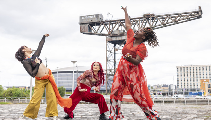 Three dancers from Whacking Scotland strike dynamic poses on a cobblestone plaza in Glasgow, with the Finnieston Crane and SEC Armadillo behind them. On the left, Nicolette McCormack leans back with an arm raised, wearing yellow trousers and a black mesh top. In the center, Leah James strikes a low stance in red velvet trousers, her long red hair down. On the right, Dorine Mwesiga reaches upward mid-motion, wearing a flowing, semi-transparent red dress over boldly patterned wide-leg pants.
