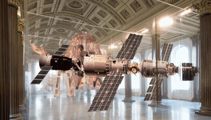 Large transparent foil sheets hang in rows from between neoclassical columns inside an ornate hall with a decorative ceiling. Different Sci-fi mages are printed on each row. On the first you can see a large spacecraft with multiple solar panels. With other cyborg images behind it from films like Blade Runner and Alien. The scene blends futuristic technology with a grand, museum‑like architectural setting.