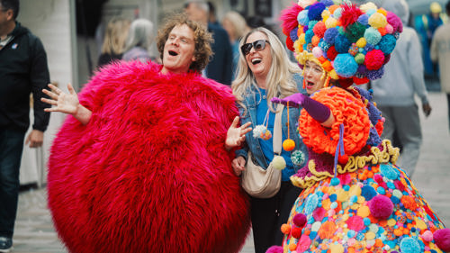 Two performers interacting with a smiling member of the public on a city street in daylight. One is wearing a large, fluffy, round costume. The other a dress, hat and umbrella covered in colourful pom poms.