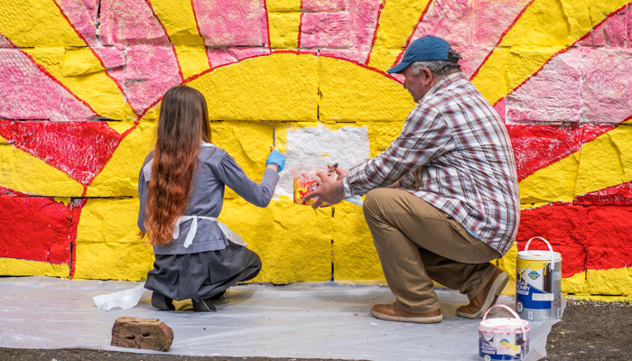 A child and adult paint a mural on a wall. It shows a setting sun, it is bright yellow and pink.