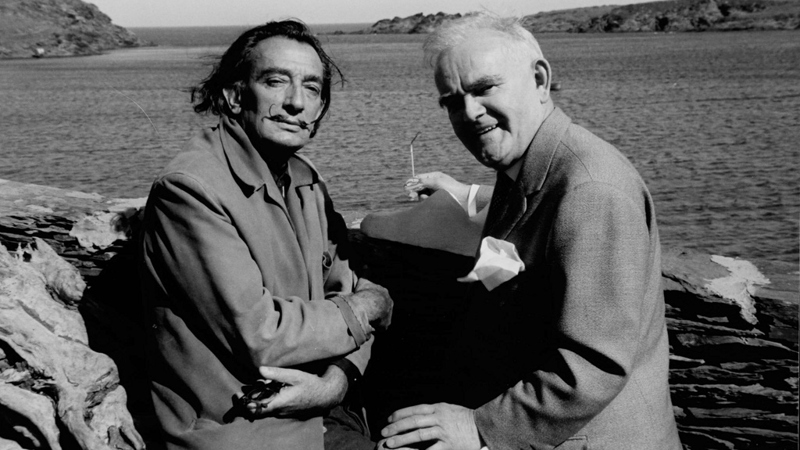Black-and-white photograph of two men standing side by side at a stone seawall overlooking a calm coastal inlet with rocky islands in the background. The man on the left has dark, shoulder-length hair and a thin mustache, with a cigarette or cigarillo in his mouth, while the man on the right has short white hair and smiles toward the camera, holding a pair of eyeglasses. Both wear light-colored jackets, and the scene appears to have been taken on a bright day by the water.
