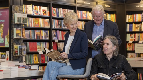 Three people are looking at books together in a bookshop. They are smiling at each other and each has a book in their hands. Two of them are sitting down, with one standing behind.